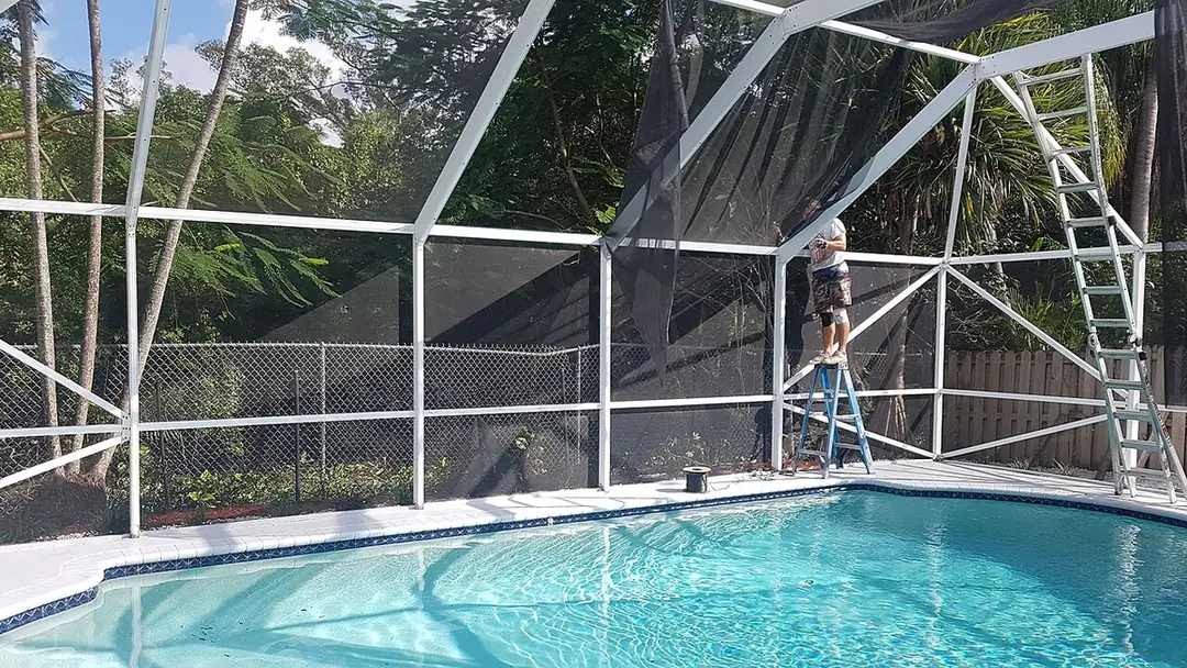 Worker on a stepladder replacing screen panels inside a white-framed pool cage