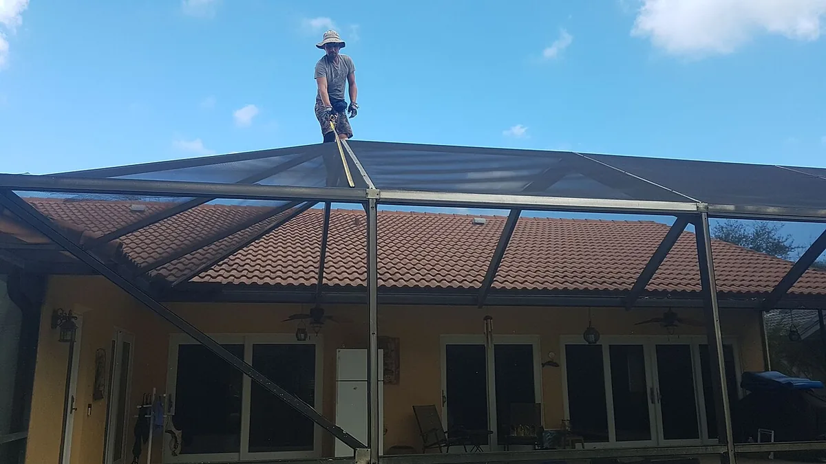 Worker standing on a dark pool cage roof beam above a tile-roofed house